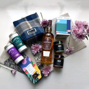 Mothers day products with bottle of whisky, chocolate, whisky teas and honeys set on a grey background with pink flowers.