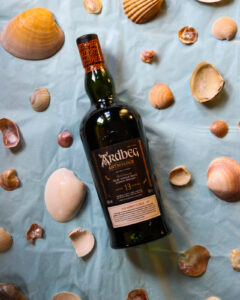 Bottle of Ardbeg Anthology, The Harpy's Tale 13 year old on a light blue background, surrounded by sea shells.