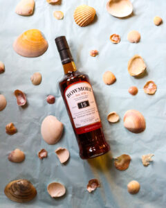 Bottle of Bowmore 15 year old pictured on a light blue background, surrounded by sea shells.