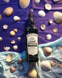 Bottle of Cutty Sark Prohibition pictured on a two toned, dark and light blue background, surrounded by sea shells.