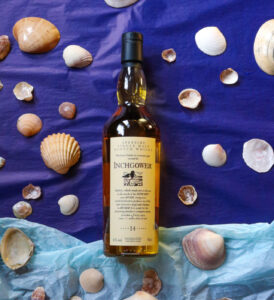 Bottle of Inchgower 14 year old pictured on a two toned, dark and light blue background, surrounded by sea shells.
