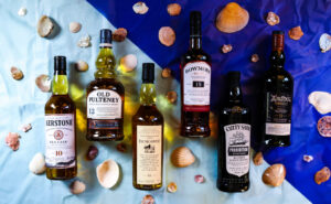Six bottles of whisky pictured on a two toned light and dark blue background, surrounded by sea shells.