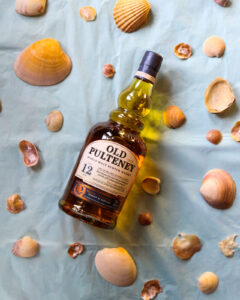 Bottle o Old Pulteney 12 year old pictured on a light blue background, surrounded by sea shells.