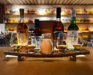 A whisky flight with a water jug in the middle is placed on a bar with whisky bottles with the matching bottles of whisky.