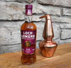 Bottle of Loch Lomond 14 Scotch Whisky with a mini copper pot still against a stone backdrop.