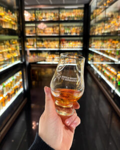 Dram of Scotch whisky against a glass cabinet collection of 3,384 whiskies.