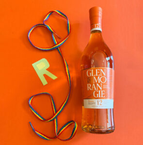 Bottle of Glenmorangie against an orange background with rainbow laces and the letter R.