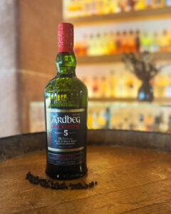 Bottle of Ardbeg Wee Beastie on a barrel accompanied with peppercorns with bar in the background.