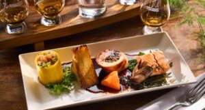 A Taste of Scotland meal and flight at Amber Restaurant at The Scotch Whisky Experience. The plate features a fish and chicken dish accompanied by vegetables, garnished with herbs, and a selection of sauce, placed on a wooden table.