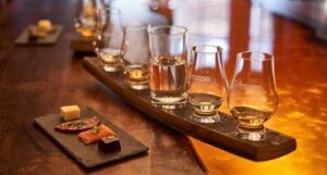 Whisky tasting flight along with food samples on an amber table at The Scotch Whisky Experience. The food highlights include cheese, venison salami, smoked salmon and a square of dark chocolate.
