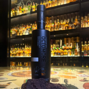 Bottle of Octomore 16.1 Scotch whisky with peices of peat, placed on a table against a collection of Scotch whisky in glass cabinets.