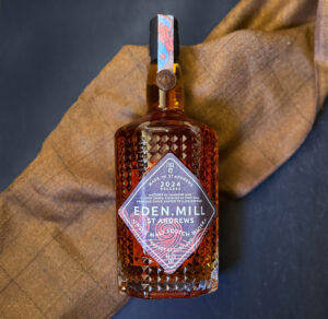 Bottle of Eden Mill St Andrews 2024 Scotch whisky on on top of a brown tartan scarf against a black backdrop.