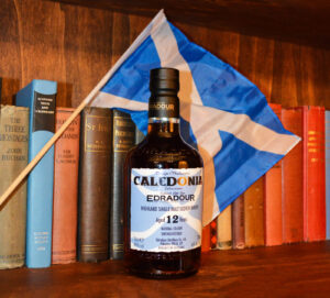 Bottle of Edradour Caledonia Scotch whisky with a Scotish flag against a bookshelf.