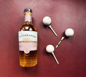 Bottle of Kingsbarns Doocot Scotch whisky with golf balls and pins against a maroon background.