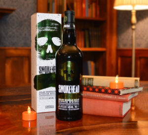 Bottle of Smokehead Unfiltered Scotch whisky and box on a amber table accompanied with books and candles with a bookcase in the background.