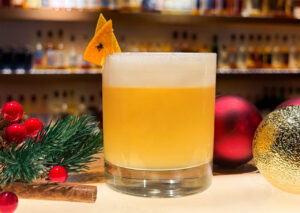 Photo of a whisky sour in front of a whisky bar. The cocktail is in a short glass and is orange/yellow in colour with foam. It is garnished with orange peel and cloves. Surrounding the glass is pine, berries and Christmas baubles for a festive feel.