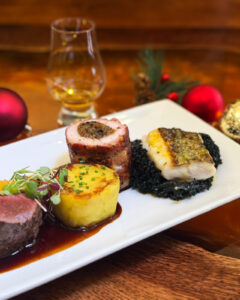 Image of a trio main with chicken, fish and lamb accompanied with crispy potato. sauce and garnishes. The plate is placed on an amber coloured table with a dram of whisky, decorated with Christmas baubles.