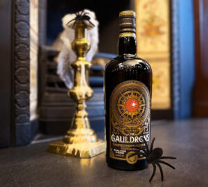 Bottle of The Gauldrons Scotch whisky next to a fireplace with a gothic candle, spider and spider webs.