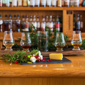 Image of a whisky flight with festive decorations such as pine and berries accompanied with a slice of cheese. There is a whisky bar in the background.