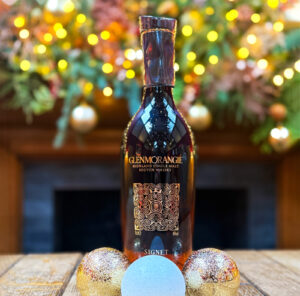 Bottle of Glenmorangie Signet Scotch whisky on top of a whisky barrel with Christmas baubles. There is festive foliage and decorations in the background.