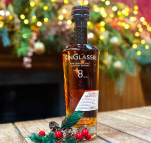 Image of KinGlassie Double Matured Scotch whisky on a whisky barrel. There is winter foliage and lights in the background and holly next to the bottle.