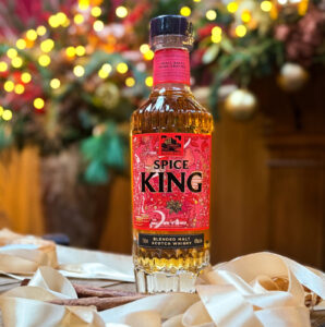 Bottle of Spice King Scotch whisky on a whisky barrel with gold ribbon and cinnamon sticks. There is festive foliage and decorations in the background.