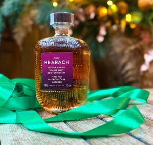Bottle of The Hearach Oloroso Scotch whisky on top of a whisky barrel with green ribbon. There is festive foliage in the background.