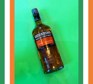 Bottle of Auchentoshan American Oak Scotch whisky on a green background with a border of Irish flags.