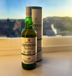 Bottle of Laphroaig 10 with whisky box on top of a white surface. There is blue skies and part of Edinburgh's skyline in the background.