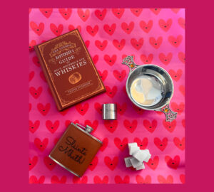 Image of a whisky book, quaich, hipflask, measure and whisky stones on top of pink wrapping paper with love hearts.