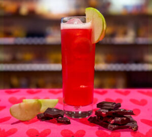Photo of a Cherry Orchard cocktail which shows pink liquid in a tall glass with an apple slice. The drink is placed on pink love heart wrapping paper and is accompanied with dark chocolate and apple slices. In the background is a blurred whisky bar.