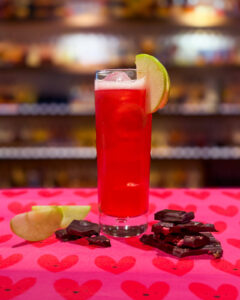 Photo of a Cherry Orchard cocktail which shows pink liquid in a tall glass with an apple slice. The drink is placed on pink love heart wrapping paper and is accompanied with dark chocolate and apple slices. In the background is a blurred whisky bar.