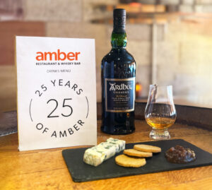 Blue cheese with oatcakes and chutney on a black slate is placed on a wooden barrel top. There is a also a bottle of Ardbeg Uigeadail Scotch whisky, a glass of whisky and a Amber Restaurant Menu highlighting its 25th anniversary.