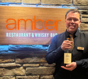 Image of restaurant manager smiling and holding a bottle of Oban 14 Scotch whisky, The male manager is standing in front of a sign that reads 'Amber Restaurant & Whisky Bar'