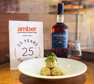 Three haggis bon bons with garnishes on a white plate is placed on a wooden barrel top. There is a also a bottle of Glendronach 18 Scotch whisky, a glass of whisky and a Amber Restaurant Menu highlighting its 25th anniversary.