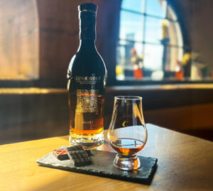 A bottle of Glenmorangie Scotch whisky is placed on a table accompanied with dark chocolate and a glass of whisky on a black slate. There is a curved window in the background.