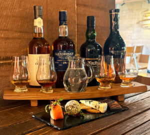 A flight of Scotch whisky is placed on a wooden table with whisky bottles in the background. In front of the flight is a slate of tapa style food.