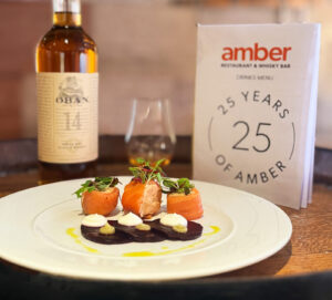 Three pieces of rolled seat trout with layered beetroot and garnishes on a white plate is placed on a wooden barrel top. There is a also a bottle of Oban 14 Scotch whisky, a glass of whisky and a Amber Restaurant Menu highlighting its 25th anniversary.