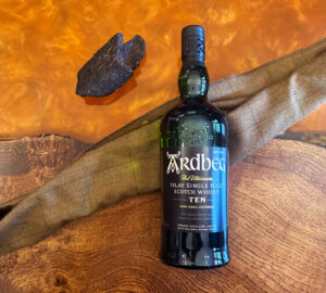 Bottle of Ardbeg Scotch whisky on an amber table with a piece of peat and a tartan sash.