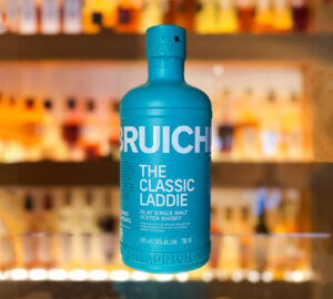 Bottle of Bruichladdich Classic Laddie Scotch whisky placed on a glass surface with an illuminated whisky bar as the backdrop.