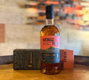 Bottle of Meikle Tòir Chinquapin Scotch whisky placed on a wooden barrel, accompanied with its whisky box. In the background is a stone wall and wooden light feature.