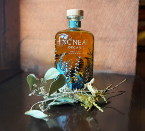 Bottle of Nc'nean Organic Scotch whisky on a black surface with a stone wall as a backdrop. The bottle is accompanied with green foliage.