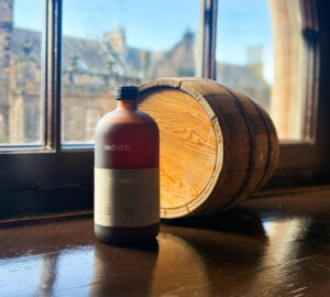 Bottle of Woven Organic Scotch whisky placed on a wooden windowsill accompanied with a miniature wooden barrel.
