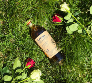 Bottle of Arran 10 Scotch whisky sat upon grass. There is 1 red and white rose placed on either side. The sun is shining down on the bottle.