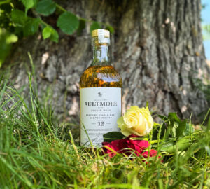Bottle of Aultmore 12 Scotch Whisky placed on the grass next to a tree. On its right is pink roses and a white rose piled up.