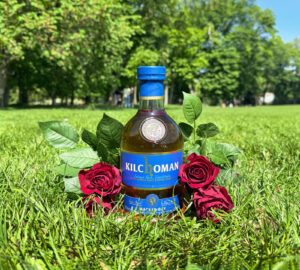 Bottle of Kilchoman Machir Bay Scotch whisky placed on grass with red roses. There is a park and trees in the background.