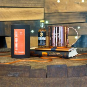 Image of corporate gifts on top of a whisky barrel with a wooden map of Scotland in the background. The gifts include whisky tea, a minature whiksy, a copper mug, and a tasting notes book.