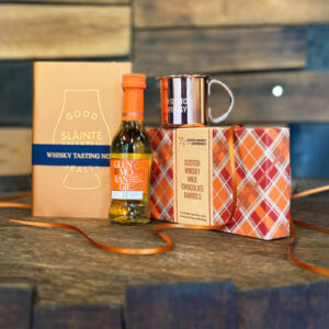 Image of corporate gifts on top of a whisky barrel with a wooden map of Scotland in the background. The gifts include a tasting notes book, a copper measure, chocolate and a minature whisky.