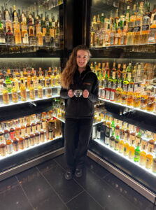 Female staff member of The Scotch Whisky Experience standing in front of a whisky collection. She is holding a quaich and smiling to the camera.