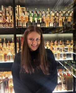 Photo of female staff member siling to the camera with a lit up whisky collection in the background.